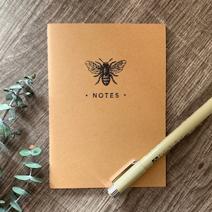 May include: A brown notebook with a bee illustration and the word "NOTES" printed on the front cover. A beige pen is lying on top of the notebook.
