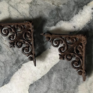 May include: Two ornate, cast iron shelf brackets with a dark brown, aged finish. The brackets feature a decorative scrollwork design and are set against a marbled gray and white background. These brackets are ideal for adding a vintage touch to any space.