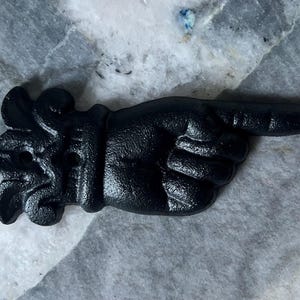 May include: A black cast iron hand pointing to the right. The hand is detailed with visible knuckles and a textured surface. The decorative piece has an ornate floral design at the base.