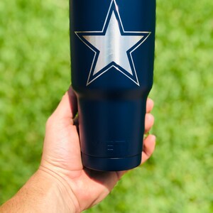 May include: A navy blue Yeti tumbler with a silver star on the front. The tumbler is being held in a hand.