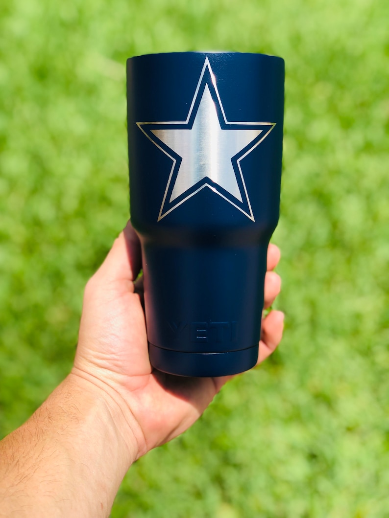 May include: A navy blue Yeti tumbler with a silver star on the front. The tumbler is being held in a hand.