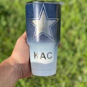 May include: A white and blue ombre tumbler with a silver star and the name "KAC" on the side. The tumbler is made by YETI.