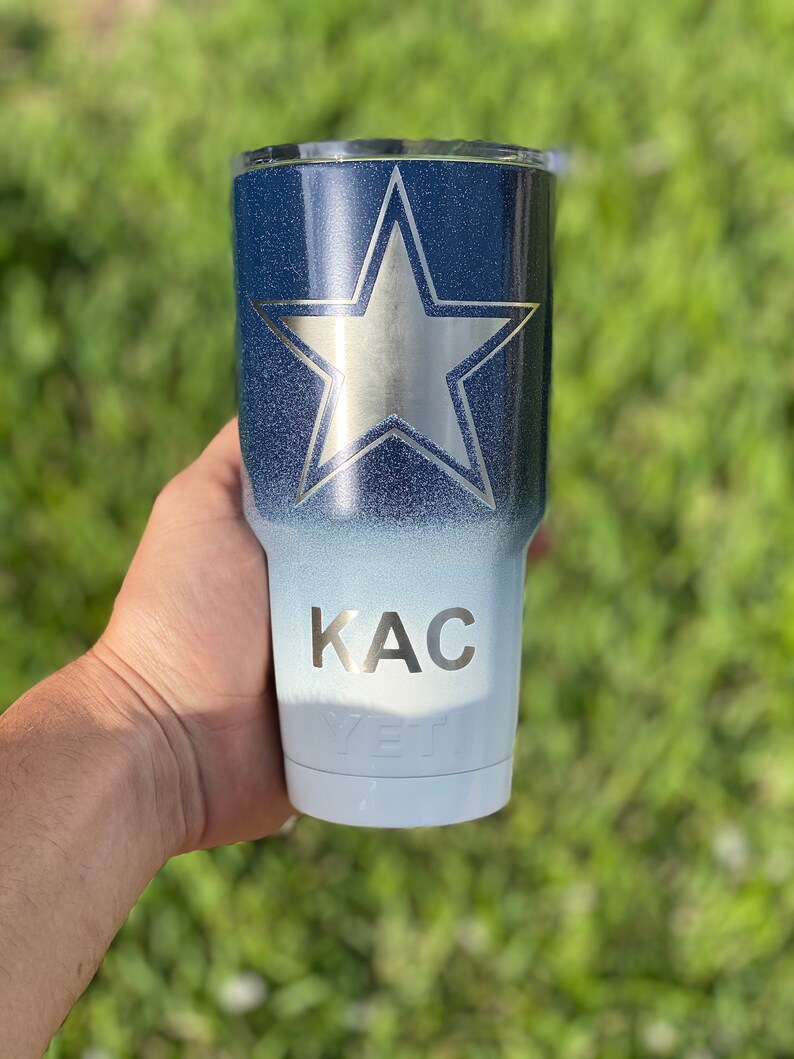 May include: A white and blue ombre tumbler with a silver star and the name "KAC" on the side. The tumbler is made by YETI.