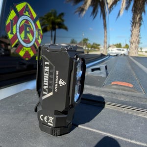 May include: Black LED flashlight with the text "LADDER 1" and a serial number. The flashlight has a clip and two bright lights. The background includes a firefighter emblem and palm trees.