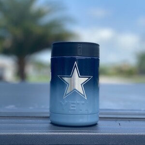 May include: A blue and white Yeti Rambler 12 oz. can cooler with a silver star design. The cooler is sitting on a blue surface.
