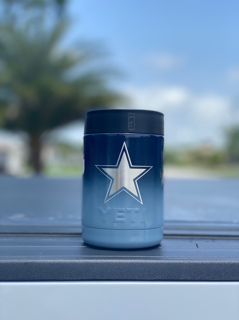 May include: A blue and white Yeti Rambler 12 oz. can cooler with a silver star design. The cooler is sitting on a blue surface.
