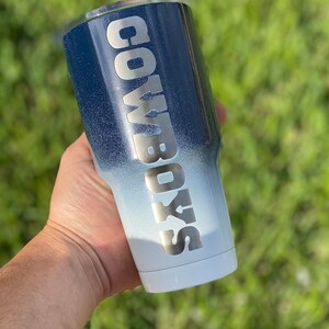 May include: A white and blue ombre tumbler with the word "COWBOYS" in silver lettering.