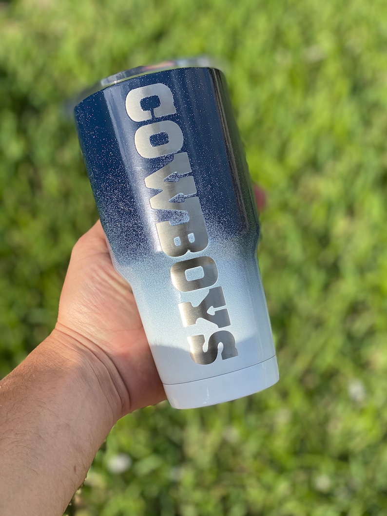 May include: A white and blue ombre tumbler with the word "COWBOYS" in silver lettering.