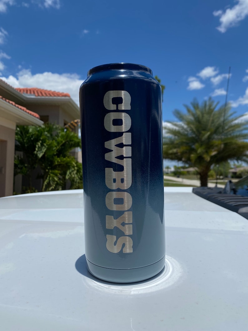 May include: A blue and black can cooler with the text "COWBOYS" in silver lettering.