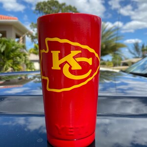 May include: Red Yeti tumbler with a yellow Kansas City Chiefs logo. The logo is a stylized arrowhead with the letters "KC" inside.