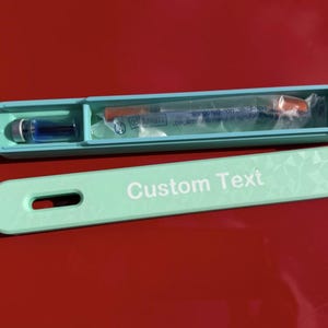 May include: A light teal medical device case with a matching lid. The case contains a vial of blue liquid and a pre-filled syringe. The lid has the words "Custom Text" in white. The case is on a red surface.