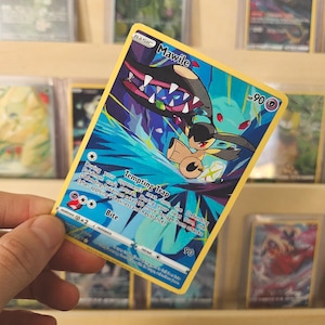 May include: A Pokémon trading card featuring Mawile, a creature with a black head and large jaws. The card has a yellow border and blue and purple background. Text includes "Mawile", "Tempting Trap", and "Bite". The card is held by a person.