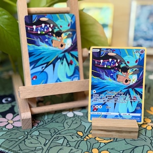 May include: A Pokémon trading card featuring Mawile with a blue and purple background. The card is displayed on a small wooden easel and a wooden block. The card's text includes "Tempting Trap" and "Bite".