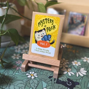 May include: A tan paper envelope labeled Mystery Pack Vol. 1 with a cartoon cat holding three blue question mark cards. The text reads 4 Alter Prints and 1 Holo Alter Print. The envelope is displayed on a small wooden easel stand.