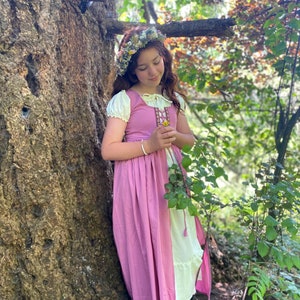 A young girl wearing a pink dress and white under dress with a floral crown stands in a forest setting. She is holding a small flower in her hand.