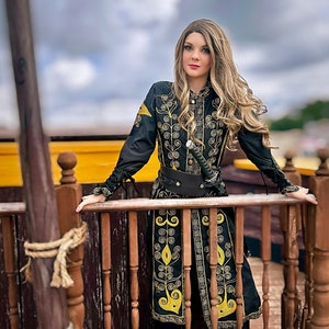 A woman in a black and gold embroidered coat stands on a wooden deck. She is holding a sword in her right hand and looking off to the side. The coat has a high collar and long sleeves. The embroidery is intricate and detailed. The woman's hair is long and blonde. She is wearing black pants and black boots. Elizabeth Swann costume from pirates of the Caribbean
