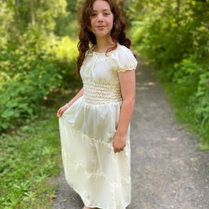 A young person wearing a white, tiered, off-the-shoulder dress with a smocked bodice. The dress has a flowy skirt and is made of a lightweight crepe fabric. The person is standing in a wooded area with green foliage in the background.