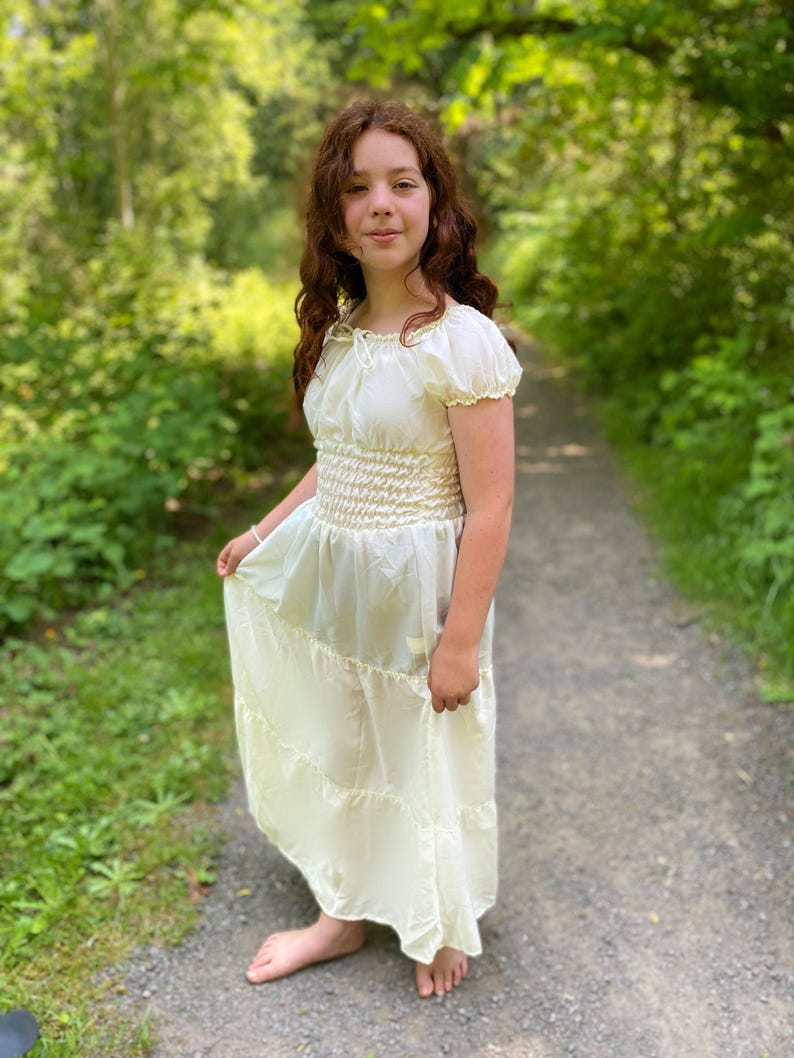 A young person wearing a white, tiered, off-the-shoulder dress with a smocked bodice. The dress has a flowy skirt and is made of a lightweight crepe fabric. The person is standing in a wooded area with green foliage in the background.