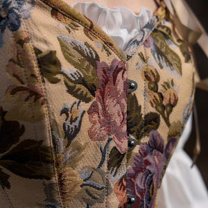 Close-up of a floral tapestry corset with a cream-colored background. The corset features a beige base with a pattern of large flowers in shades of purple, red, and green. The corset has metal clasps and is paired with a white ruffled blouse.