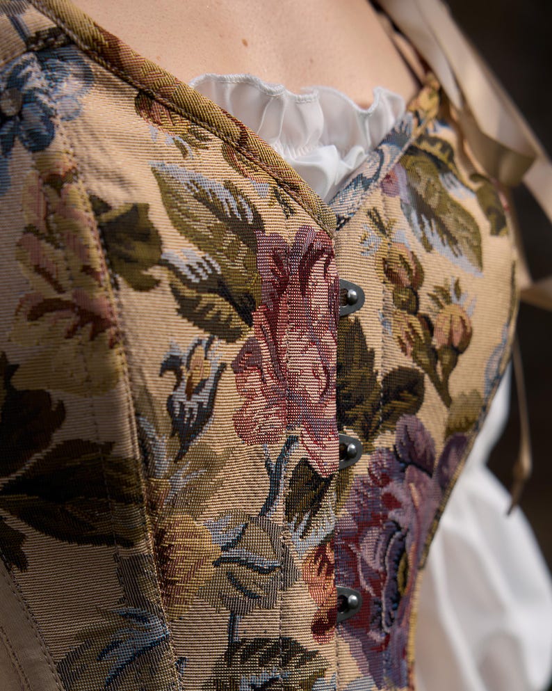 Close-up of a floral tapestry corset with a cream-colored background. The corset features a beige base with a pattern of large flowers in shades of purple, red, and green. The corset has metal clasps and is paired with a white ruffled blouse.