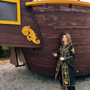 A woman wearing a black and gold embroidered costume from pirate of the Caribbean stands in front of a wooden ship. She holds a sword in her right hand.