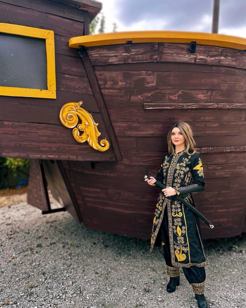 A woman wearing a black and gold embroidered costume from pirate of the Caribbean stands in front of a wooden ship. She holds a sword in her right hand.