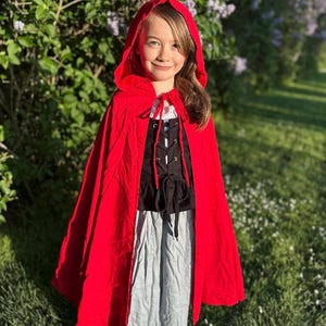 May include: A young person wearing a red hooded cape, black bodice, and gray skirt. The cape is tied at the neck with a white string. The outfit is reminiscent of a fairytale character. The background features green foliage and flowers.