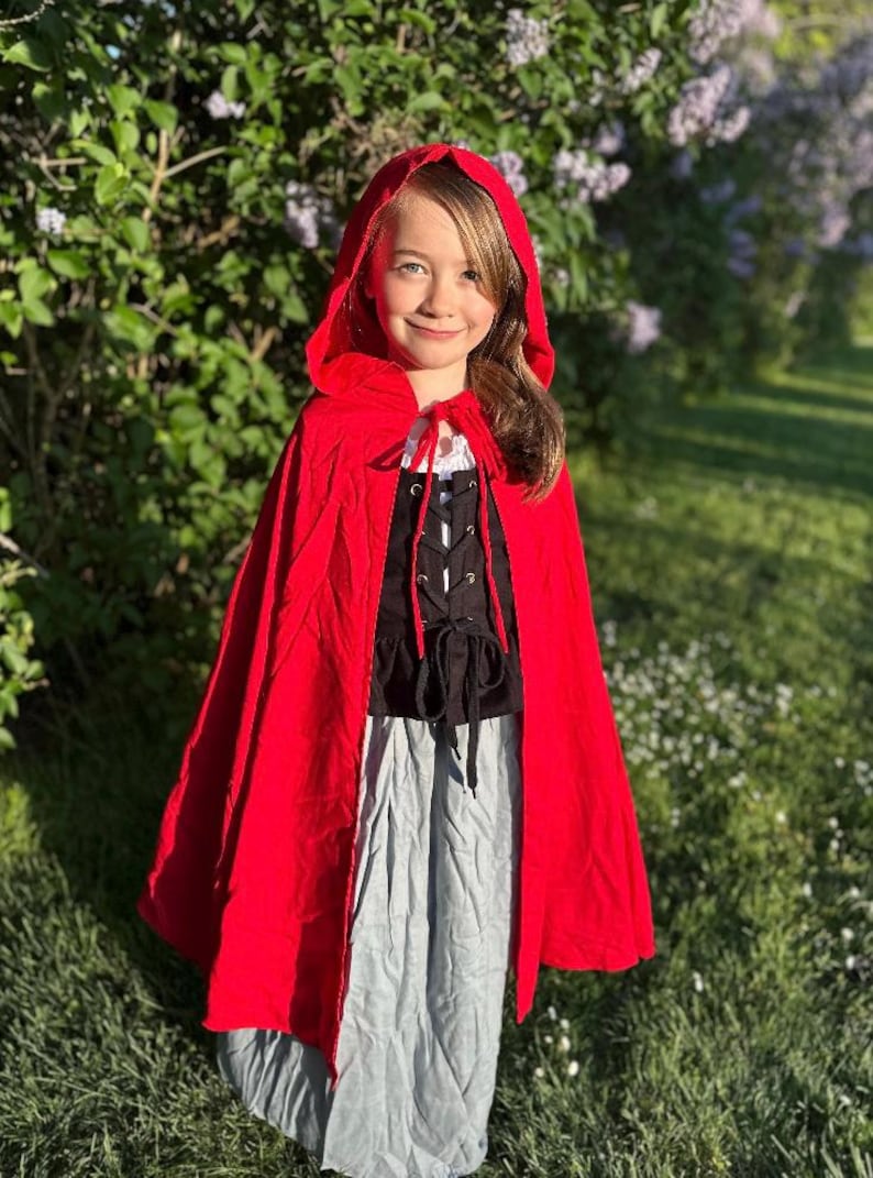 May include: A young person wearing a red hooded cape, black bodice, and gray skirt. The cape is tied at the neck with a white string. The outfit is reminiscent of a fairytale character. The background features green foliage and flowers.