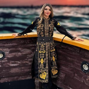 A woman wearing a black and gold embroidered coat with a black belt. She is standing on a wooden boat with a sunset in the background.