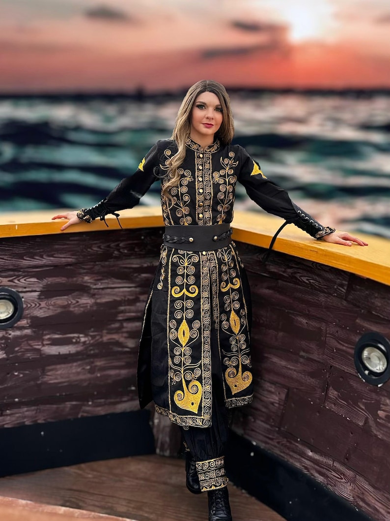 A woman wearing a black and gold embroidered coat with a black belt. She is standing on a wooden boat with a sunset in the background.