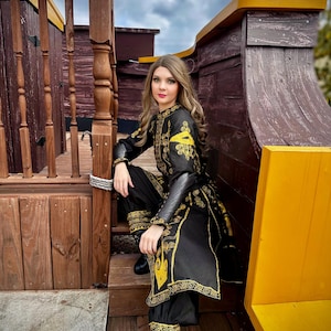 A woman wearing a black and gold embroidered costume with a long skirt and pants. She is sitting on a wooden deck with a yellow railing behind her. she is wearing an rlizabeth Swann costume