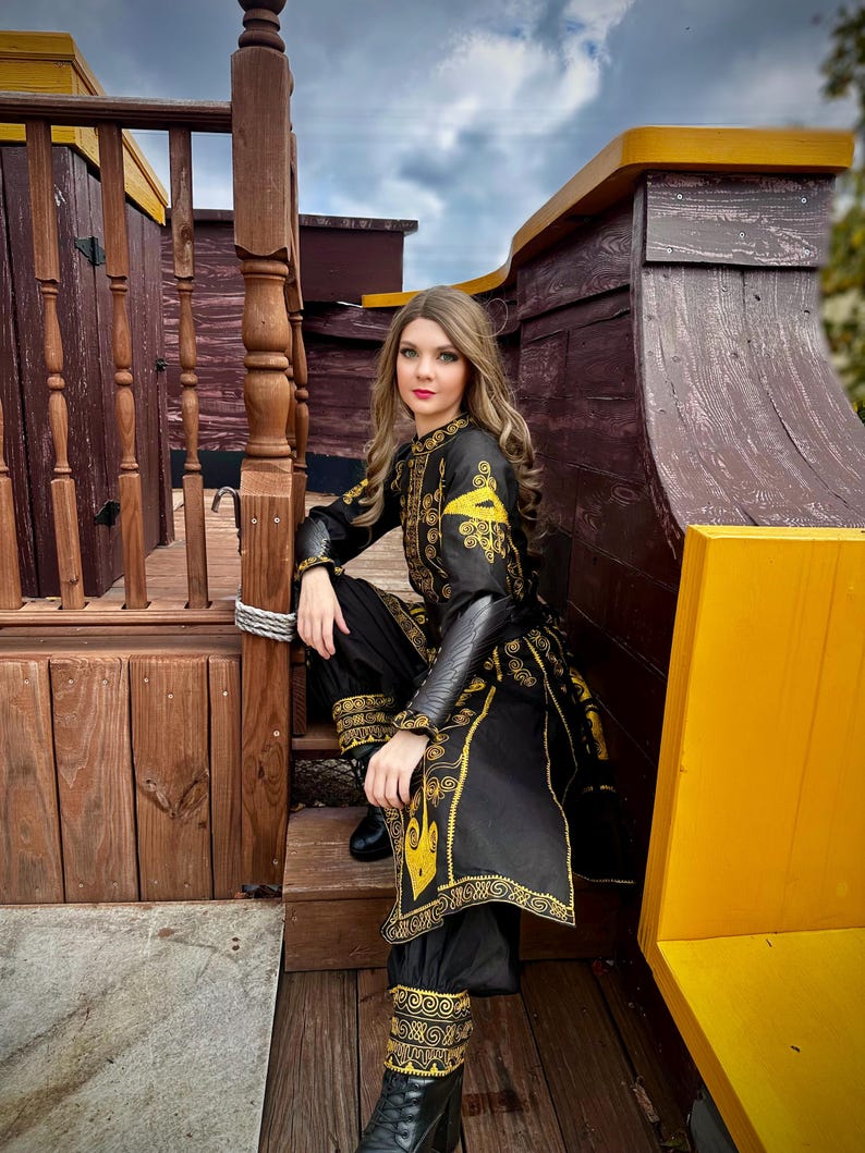 A woman wearing a black and gold embroidered costume with a long skirt and pants. She is sitting on a wooden deck with a yellow railing behind her. she is wearing an rlizabeth Swann costume