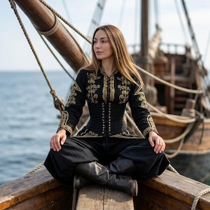 the model is sitting with her legs crossed on the bow of a pirate ship. she is wearing an Elizabeth Swann costume in black with gold embroidery.