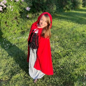 May include: A young person wearing a red hooded cape and a costume with a black bodice and a gray skirt, standing in a grassy area with flowers and trees in the background. The costume resembles a fairytale character.