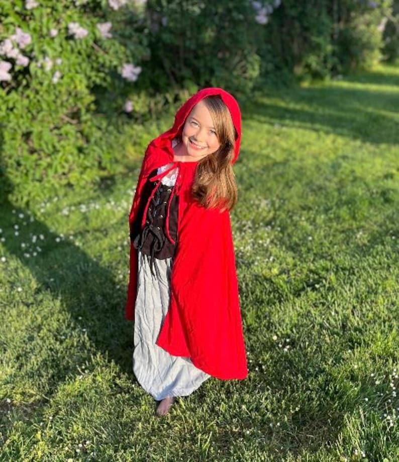May include: A young person wearing a red hooded cape and a costume with a black bodice and a gray skirt, standing in a grassy area with flowers and trees in the background. The costume resembles a fairytale character.