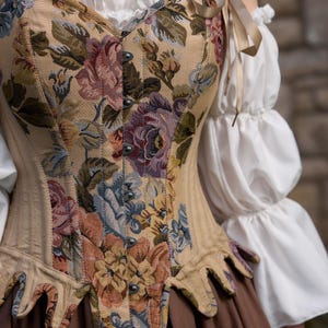 A beige corset with a floral tapestry design featuring pink, purple, and blue flowers. The corset has a pointed hemline and is paired with a white blouse with ruffled sleeves and a brown skirt. Beige ribbons are tied at the shoulders.
