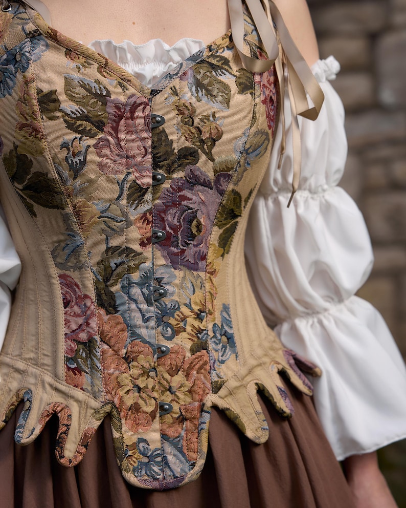 A beige corset with a floral tapestry design featuring pink, purple, and blue flowers. The corset has a pointed hemline and is paired with a white blouse with ruffled sleeves and a brown skirt. Beige ribbons are tied at the shoulders.