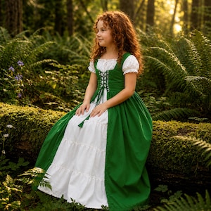 May include: A green and white dress with a corset-style bodice and short, puffed sleeves. The dress features a green skirt and a white, tiered skirt. The girl is sitting on a moss-covered log in a forest setting.