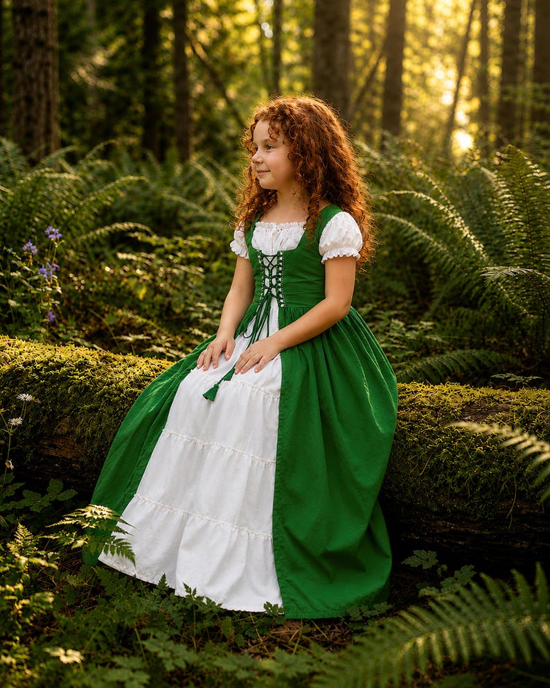 May include: A green and white dress with a corset-style bodice and short, puffed sleeves. The dress features a green skirt and a white, tiered skirt. The girl is sitting on a moss-covered log in a forest setting.