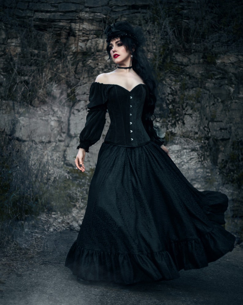 A woman in a black, off-the-shoulder veneer snd lace corset and a long, flowing lace skirt. She is wearing a black headband with a feather and has dark hair and red lipstick.