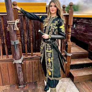 A woman wearing a black and gold embroidered jacket and matching pants with a sword at her side. She is standing on a wooden deck with a rope tied to a wooden post. Elizabeth Swann costume.