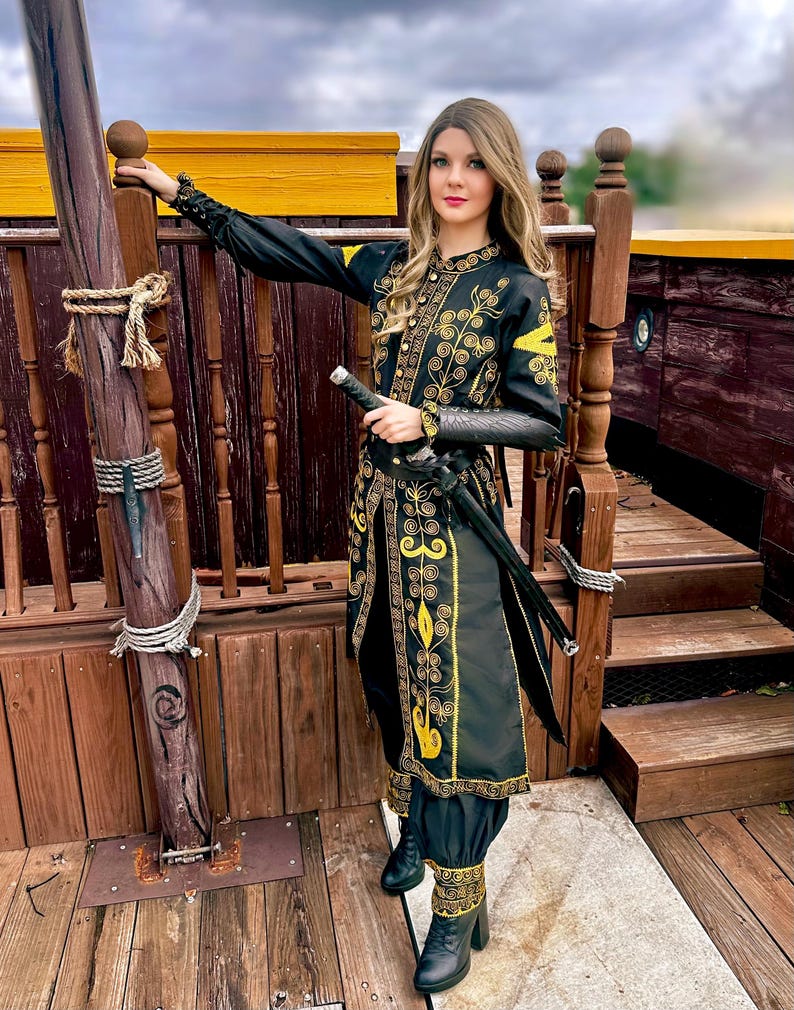 A woman wearing a black and gold embroidered jacket and matching pants with a sword at her side. She is standing on a wooden deck with a rope tied to a wooden post. Elizabeth Swann costume.