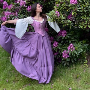 May include: A floor-length dress featuring a lavender bodice with a lace-up front and white long sleeves. The skirt is a matching lavender color with tiered layers that flare out. The neckline is trimmed with white lace, and the bodice has shoulder straps.