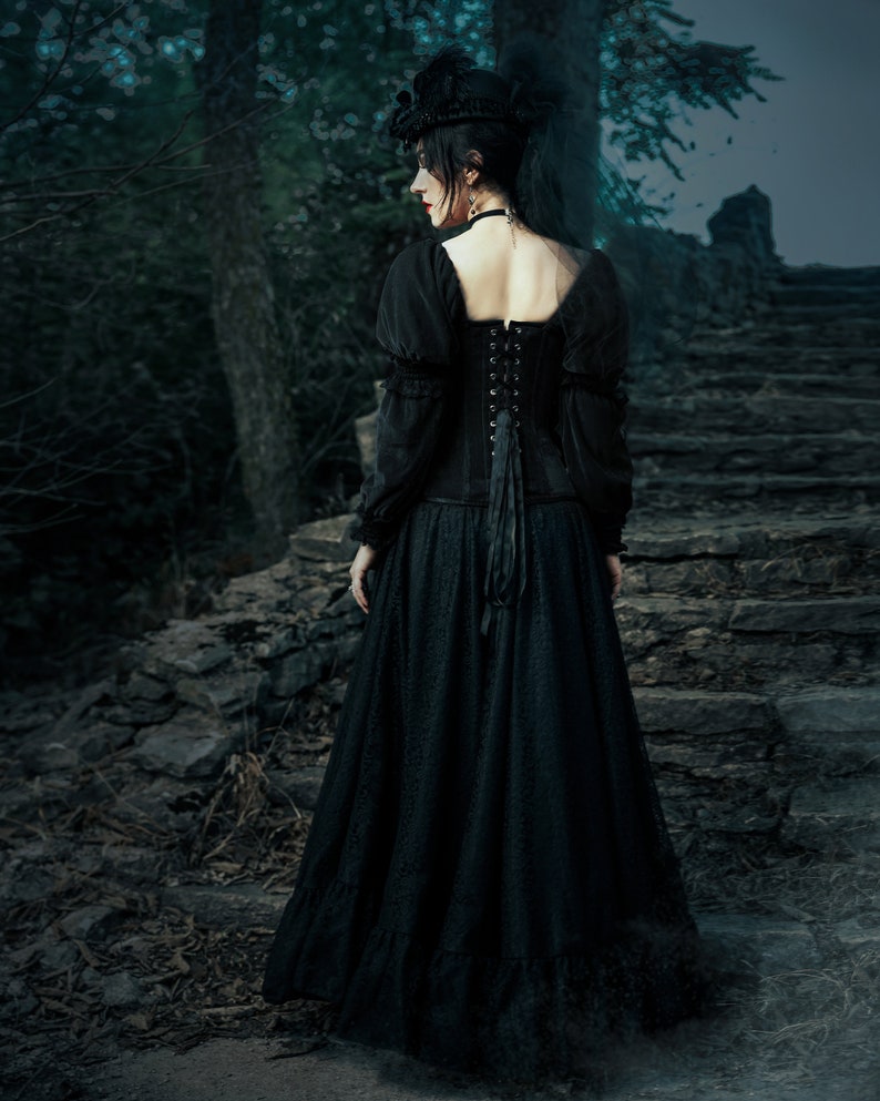 A woman in a black dress with a lace overlay and a black hat stands on a stone staircase in a dark forest setting. she is wearing a velvet and lace corset and lsve full and long skirt.