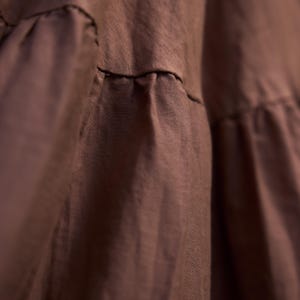 Close-up of a brown fabric garment with visible stitching and ruffled details. The fabric appears to be soft and flowing, with a slightly textured surface. The garment's design includes horizontal seams and gathered sections.
