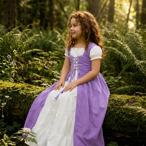 May include: A young girl with curly red hair wearing a purple and white dress with short, puffed sleeves and a laced bodice. The dress has a tiered skirt and is set against a backdrop of lush green foliage and sunlight.