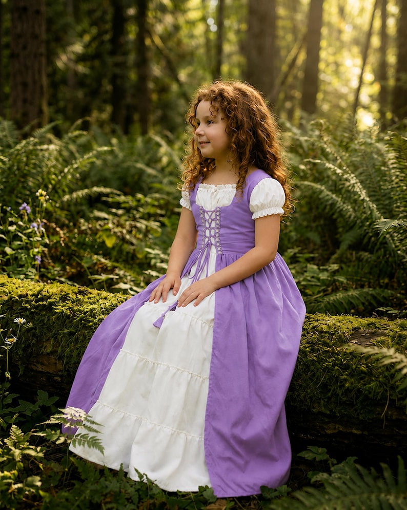 May include: A young girl with curly red hair wearing a purple and white dress with short, puffed sleeves and a laced bodice. The dress has a tiered skirt and is set against a backdrop of lush green foliage and sunlight.