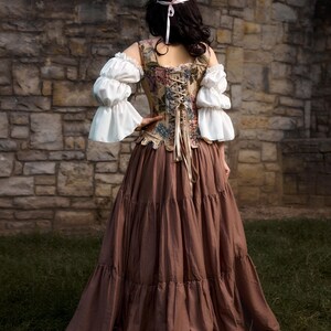A Renaissance-style dress with a floral patterned bodice, white ruffled sleeves, and a long brown tiered skirt. The back of the bodice has a lace-up design. A floral crown adorns the head.