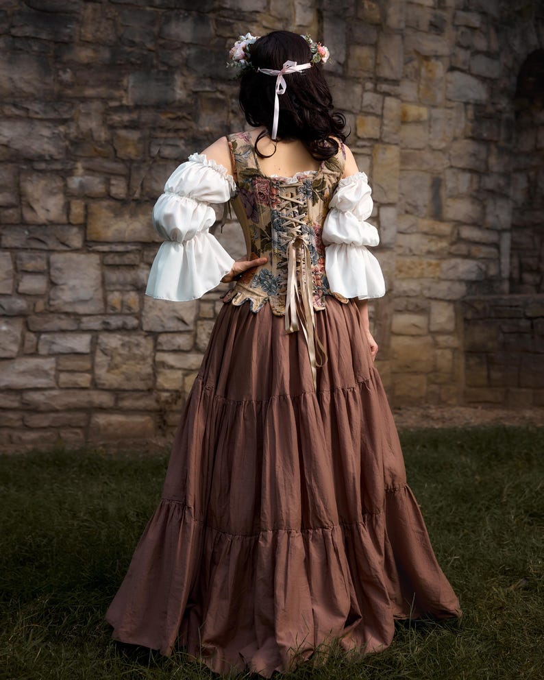 A Renaissance-style dress with a floral patterned bodice, white ruffled sleeves, and a long brown tiered skirt. The back of the bodice has a lace-up design. A floral crown adorns the head.