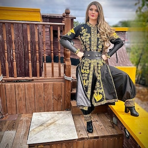 A woman wearing a black and gold embroidered costume with a long skirt and pants. She is standing on a wooden platform with a yellow railing. Elizabeth Swann from POTC.